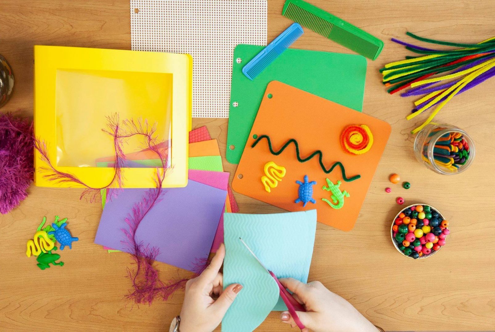 A wide variety of tactile objects on a table next to an open book. A person cuts foam with scissors.