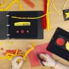 A wide variety of tactile objects in red and yellow on a table next to an open black book. A person cuts string with scissors.