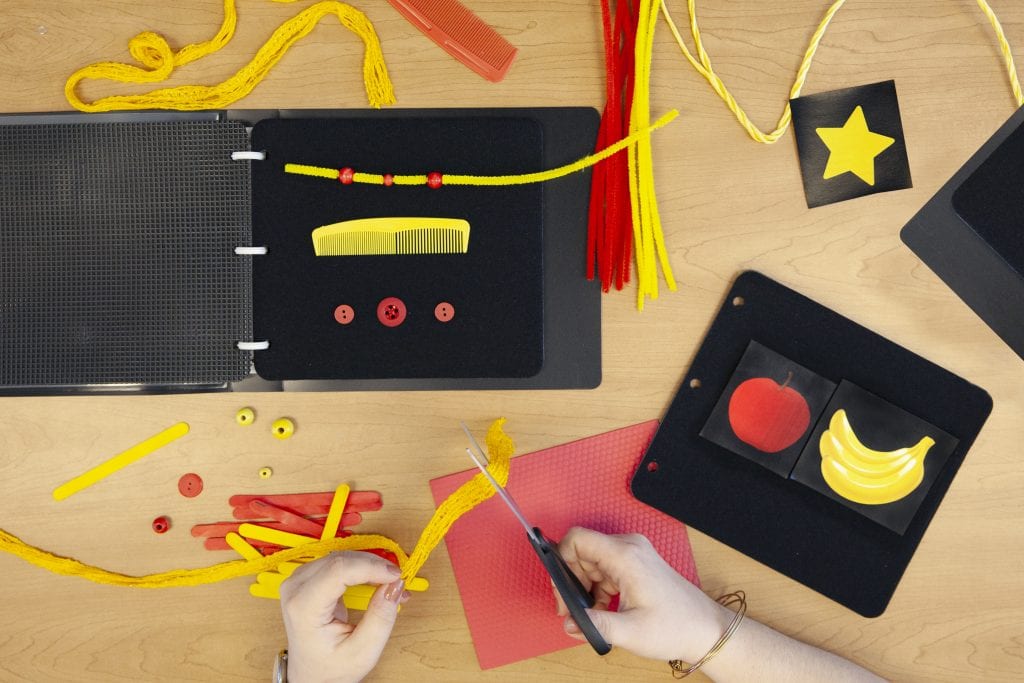 A wide variety of tactile objects in red and yellow on a table next to an open black book. A person cuts string with scissors.
