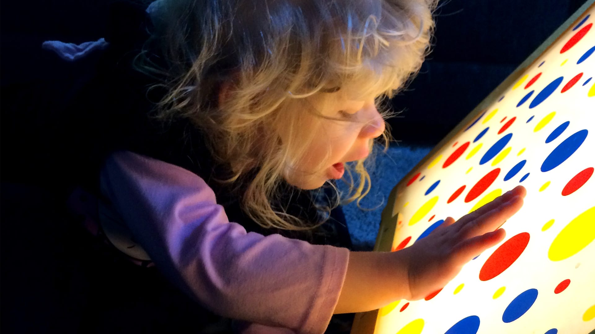 Young child playing with a light box showing red, blue, and yellow polka dots