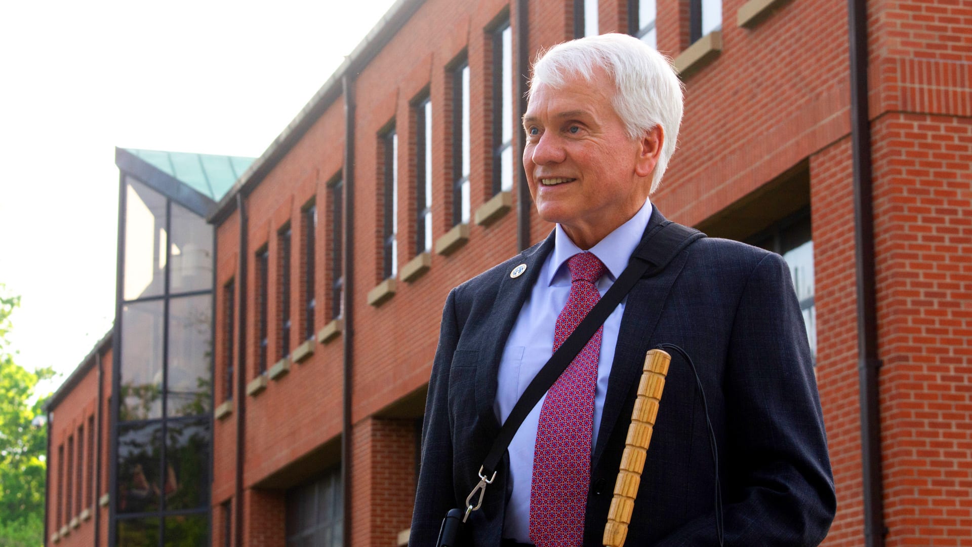 Man with refreshable braille display and white cane walking outside an office building