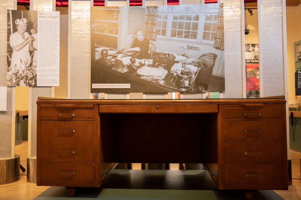 A large wooden desk, poster of Helen Keller working at that desk hangs behind