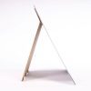 A side view of a frameless mirror propped up on a brown stand against a white background.