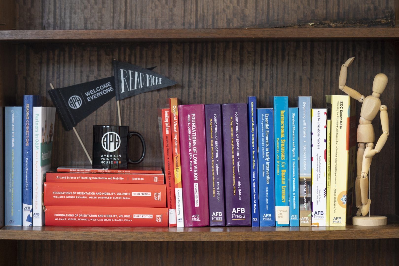 aph press books lined up on a shelf with an APH coffee mug, pennant flag, and a little wooden model figure