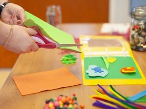 Hands cutting a piece of green art foam. On the table is an open binder with pages decorated in tactile craft elements.