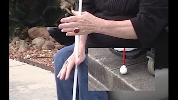 Close up of woman in wheelchair using cane to measure height of curb.