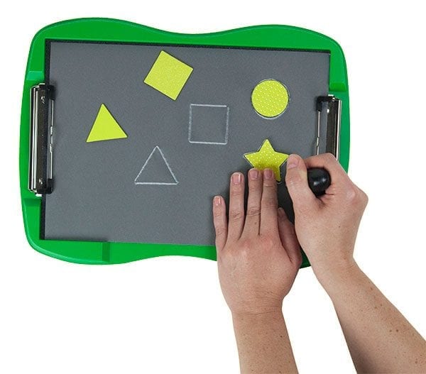 Two hands are tracing a star-shaped yellow stencil with the black stylus on tactile drawing film attached to the green TactileDoodle frame. Above the star is a circle-shaped stencil that has already been traced. To the left are two white tactile outlines of a triangle and a square next to the yellow stencils of the same shapes