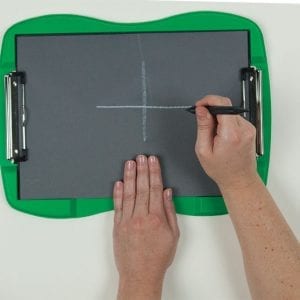 A sheet of tactile drawing film is attached to the curved green frame of the TactileDoodle. Two hands appear in the image; one is holding the film in place while the other draws a white cross in the center of the page with the black stylus