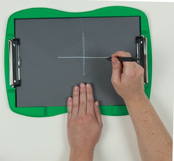 A sheet of tactile drawing film is attached to the curved green frame of the TactileDoodle. Two hands appear in the image; one is holding the film in place while the other draws a white cross in the center of the page with the black stylus