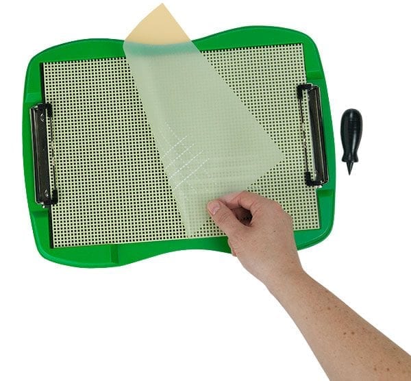 A hand is peeling back a piece of tactile drawing film from a textured crosshatch white sheet attached to the green TactileDoodle frame. The black stylus sits on the right