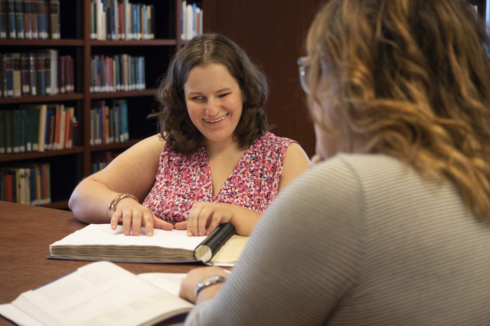 two women sitting at a table, one is reading from a braille book