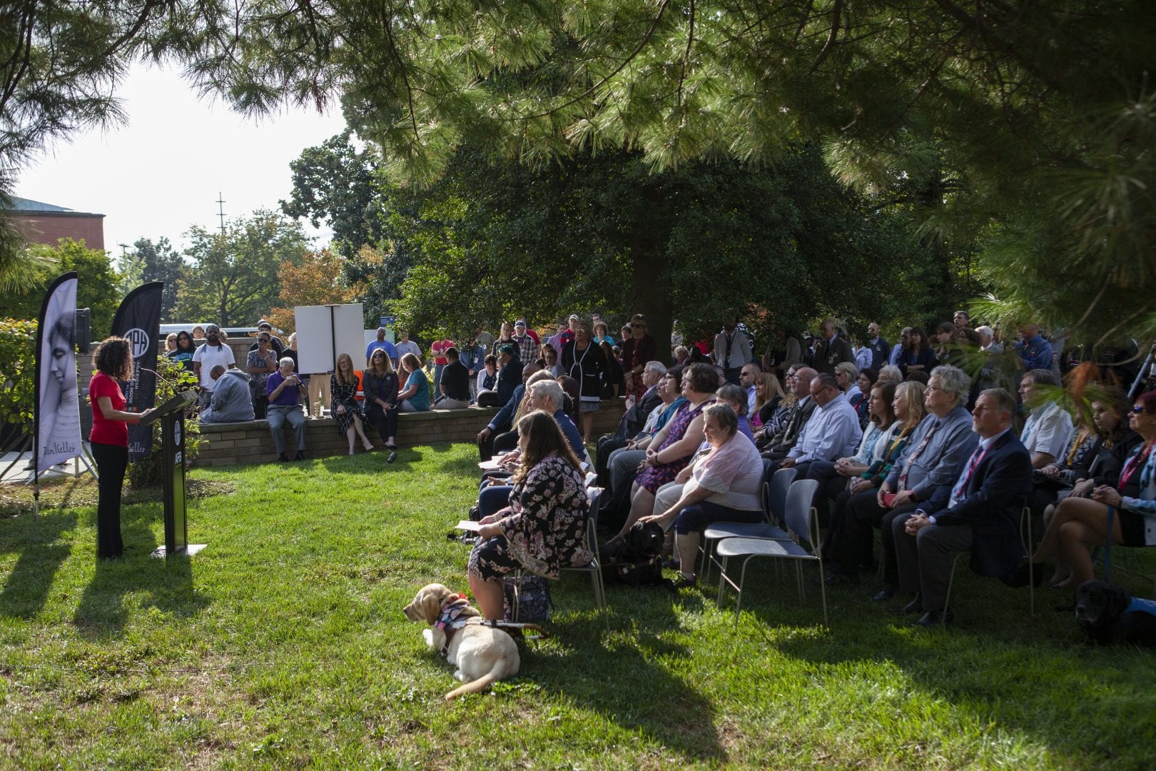 a crowd of people sitting and standing at a news conference. a woman stands at a podium.