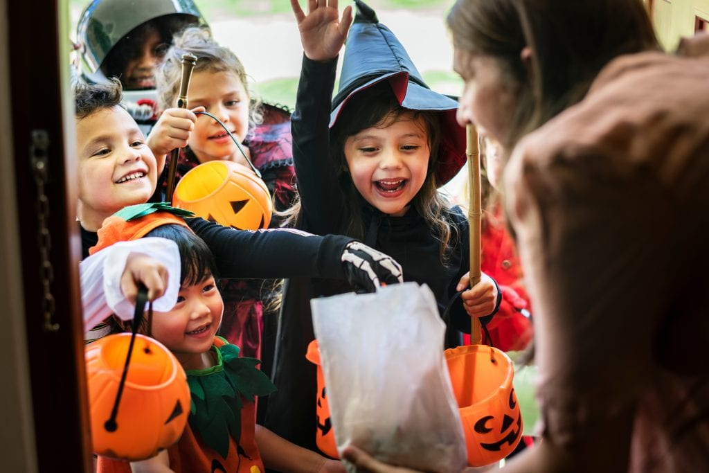 A group of kids in costume holding orange pumpkins. They're smiling and laughing as they trick-or-treat at a woman's front door.