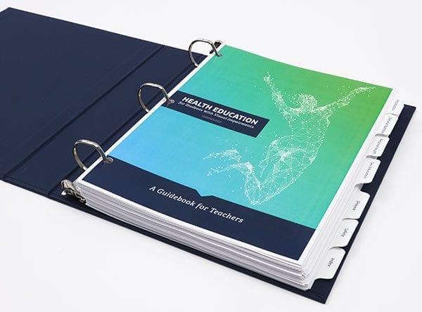 The first page of an open blue Health Education guidebook binder. Printed on the blue and green page is the title of the book and a white geometric graphic of a jumping woman. The binder contains section divider tabs.
