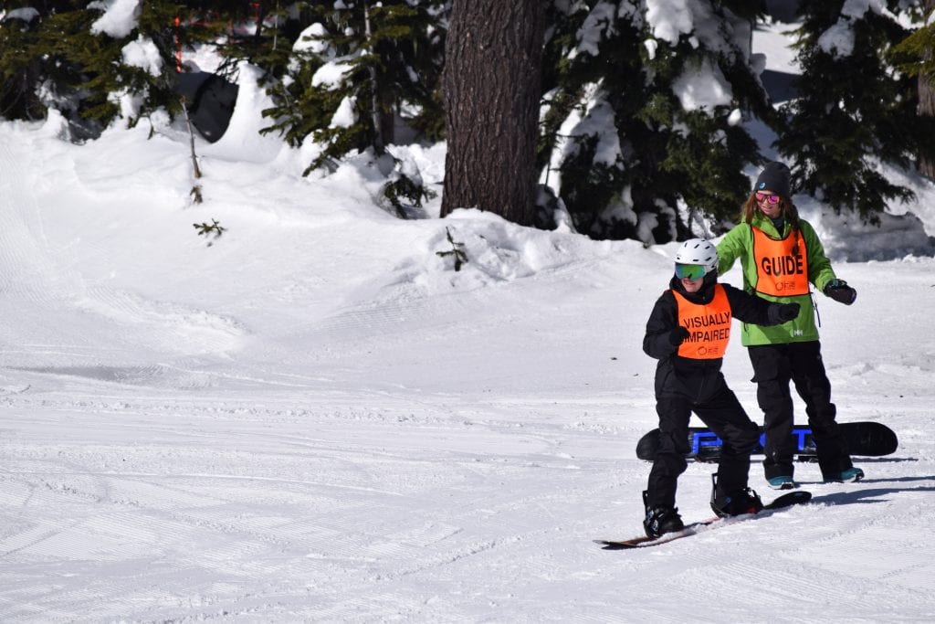 a student snowboarder in an orange vest on the snow, an instructor is behind them