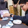 student adjusting a code jumper pod at a table covered in a network of connected pods. A tablet displays the app in the background.