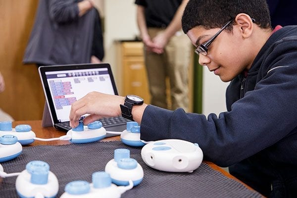 student adjusting a code jumper pod at a table covered in a network of connected pods. A tablet displays the app in the background.