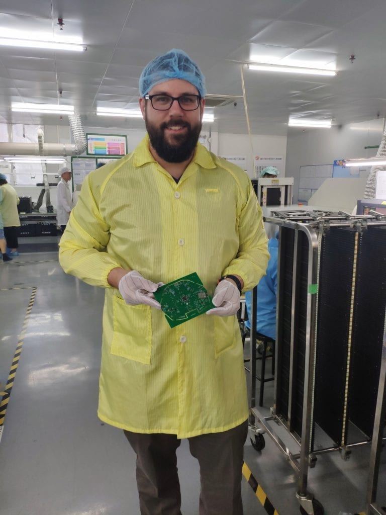 andrew in a labcoat, hair net, and rubber gloves holding a piece of circuit board and smiling