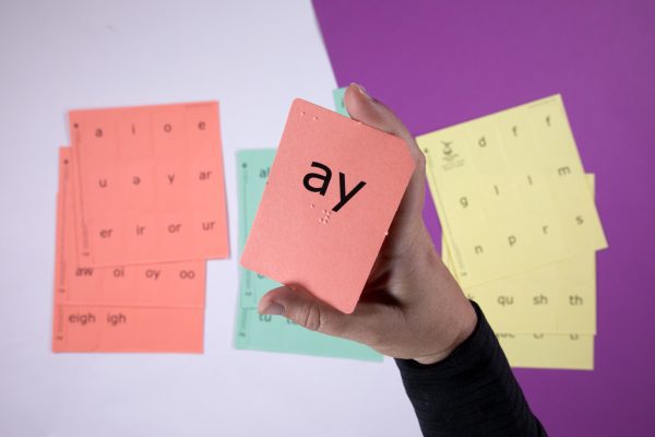 Close up of a red vowel blend sound card that reads "ay" in both print and braille. In the background are various sound card sheets.