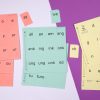 Three stacks of perforated print-braille word card sheets in red, orange, and yellow against a white and purple background.