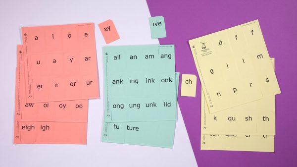 Three stacks of perforated print-braille word card sheets in red, orange, and yellow against a white and purple background.