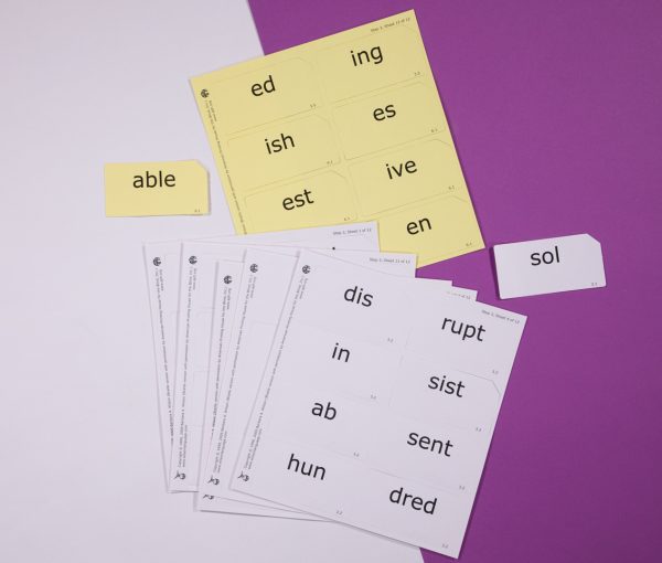 Two stacks of print-braille syllable card sheets, one yellow and one white. Next to each stack is one card that has been punched out.