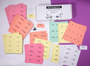 The box and full set of Wilson Reading System print-braille cards in a variety of colors laid out against a white and purple background.