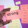 A close up of a hand holding a print-braille card with the word "strong" in both large print and braille. In the background are various perforated sound card sheets.
