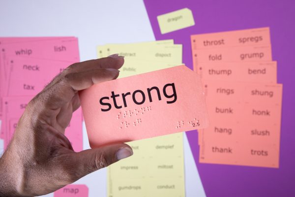 A close up of a hand holding a print-braille card with the word "strong" in both large print and braille. In the background are various perforated sound card sheets.