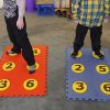 Two students standing on red and blue foam floor mat braille cells with yellow dots containing numbers.