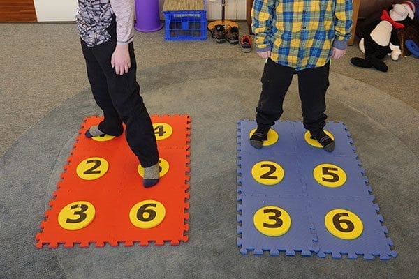 Two students standing on red and blue foam floor mat braille cells with yellow dots containing numbers.