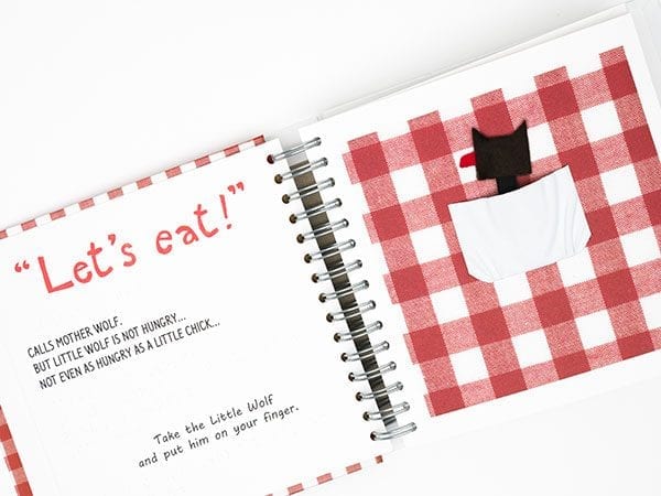An open storybook. On the left page it says "Let's eat!" in big red font. On the right page there is a red gingham background. A white pocket in the center holds a little felt figure of wolf with a red tongue.