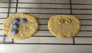 two cookies, one with circular candies positioned into a braille letter and the other with cheerios in a different braille letter format