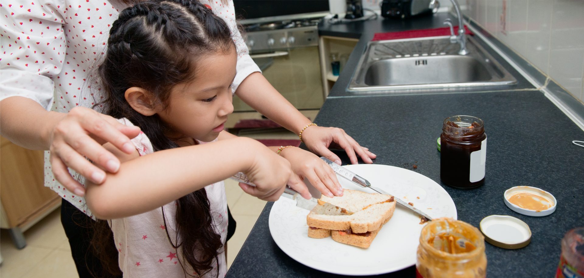 A child cutting a peanut butter sandwich with a parent’s help.