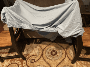 a fitted sheet stretched out over the tops of two dining room chairs, creating a covered space below