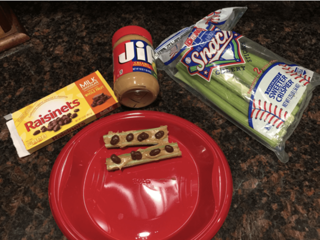 a box of raisinets, a jar of peanut butter, a bag of celery, and a plate with two assembled ants on a log treats