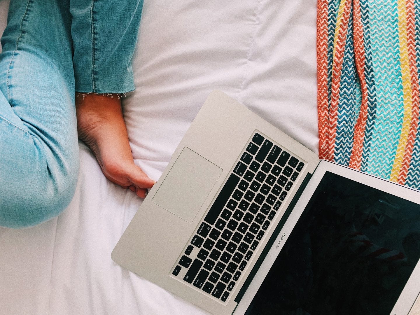 mac book and legs of someone sitting in a bed with a colorful throw blanket
