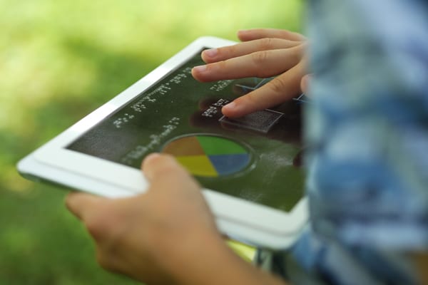 A child outside holding a tablet with an overlay. They are exploring the overlay with their finger.