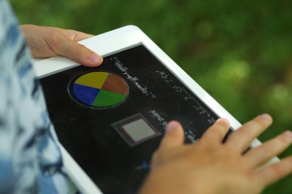 A child outside holding a tablet with an overlay. They are exploring the overlay with their finger.