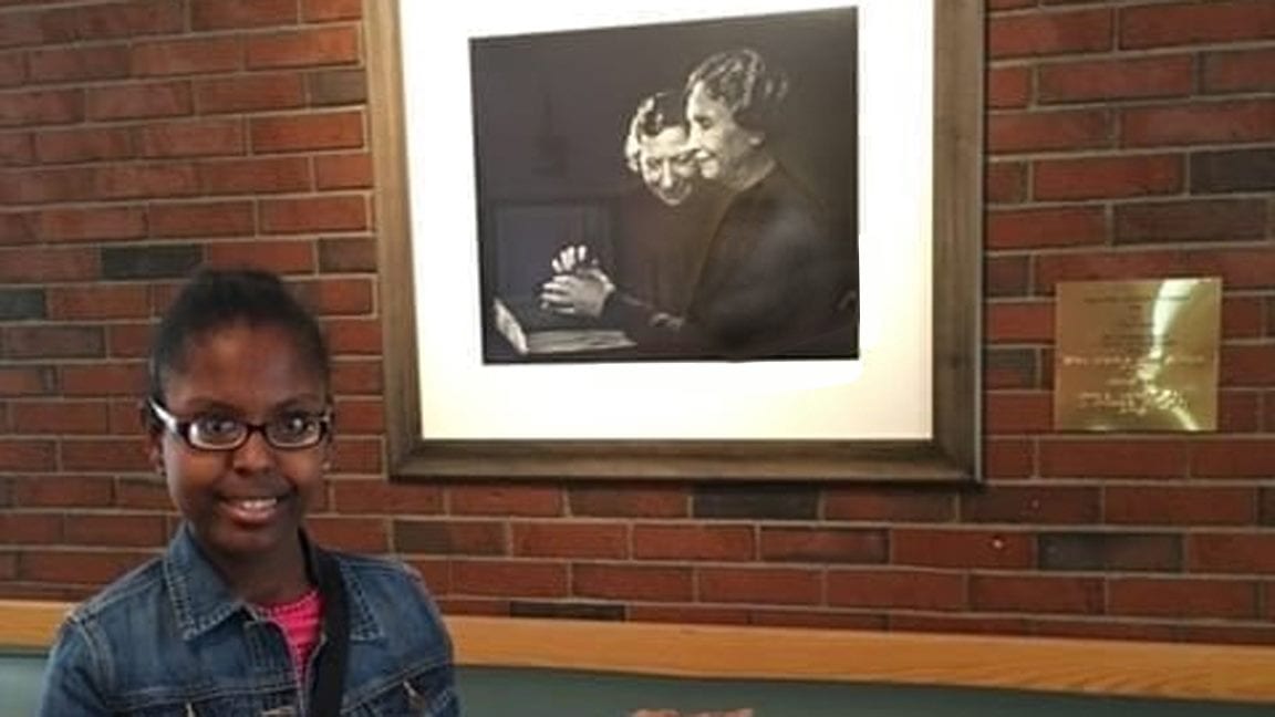 A young woman, smiling, stands in front of a framed photograph of Anne Sullivan and Helen Keller.