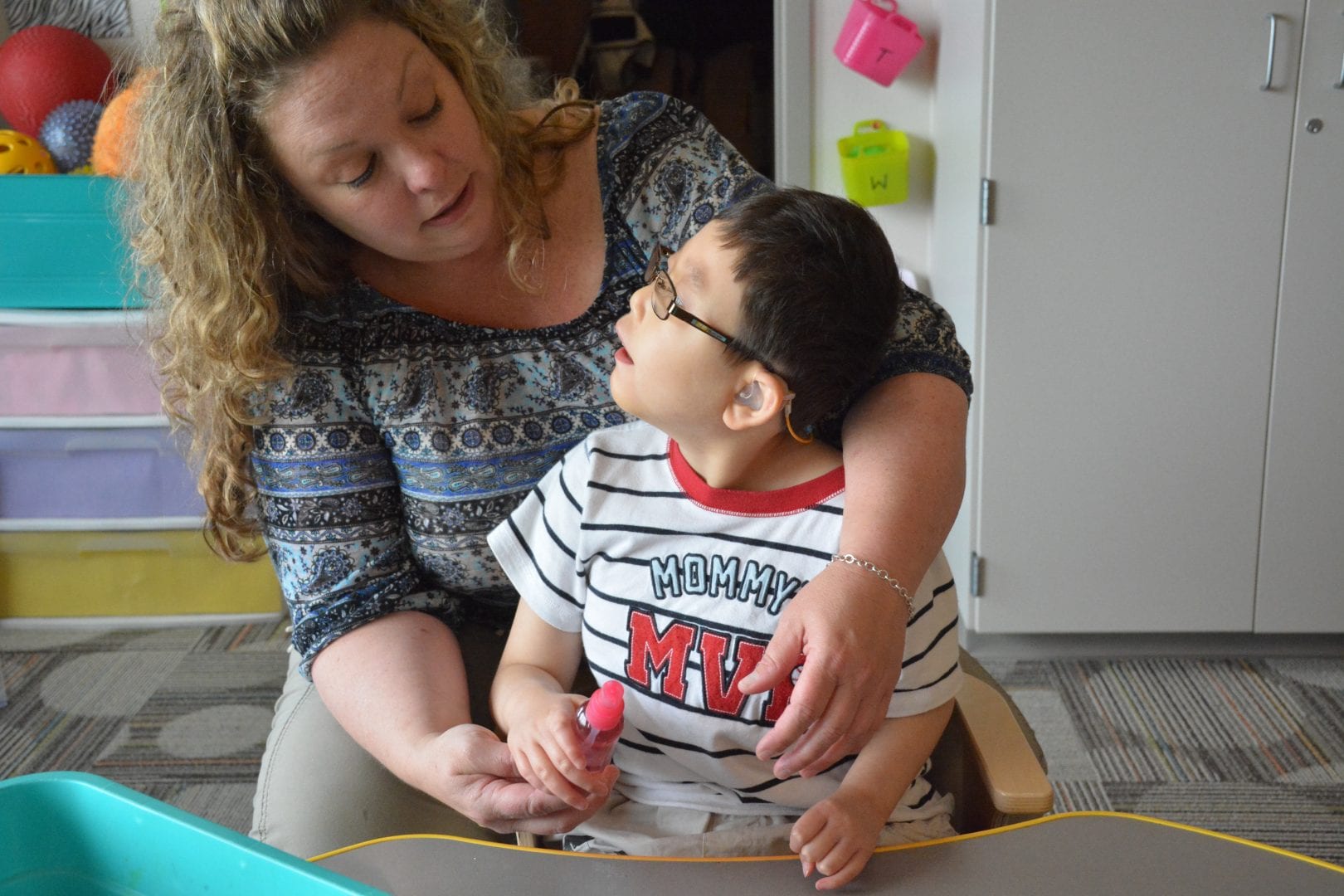 A student's teacher provides light elbow support while he grasps his water bottle and looks to her, confirming he is ready to spray his hair.