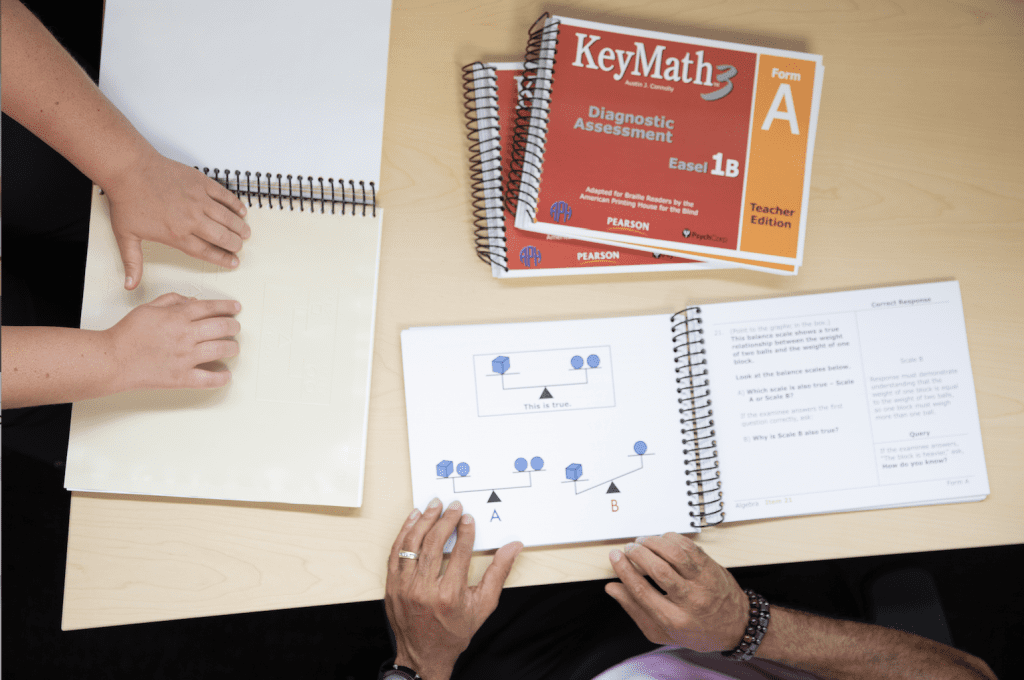 over head shot of a students hands reading braille and tactile graphics on the student book and the teacher following along with the teacher edition. The other Keymath books sit on the table.