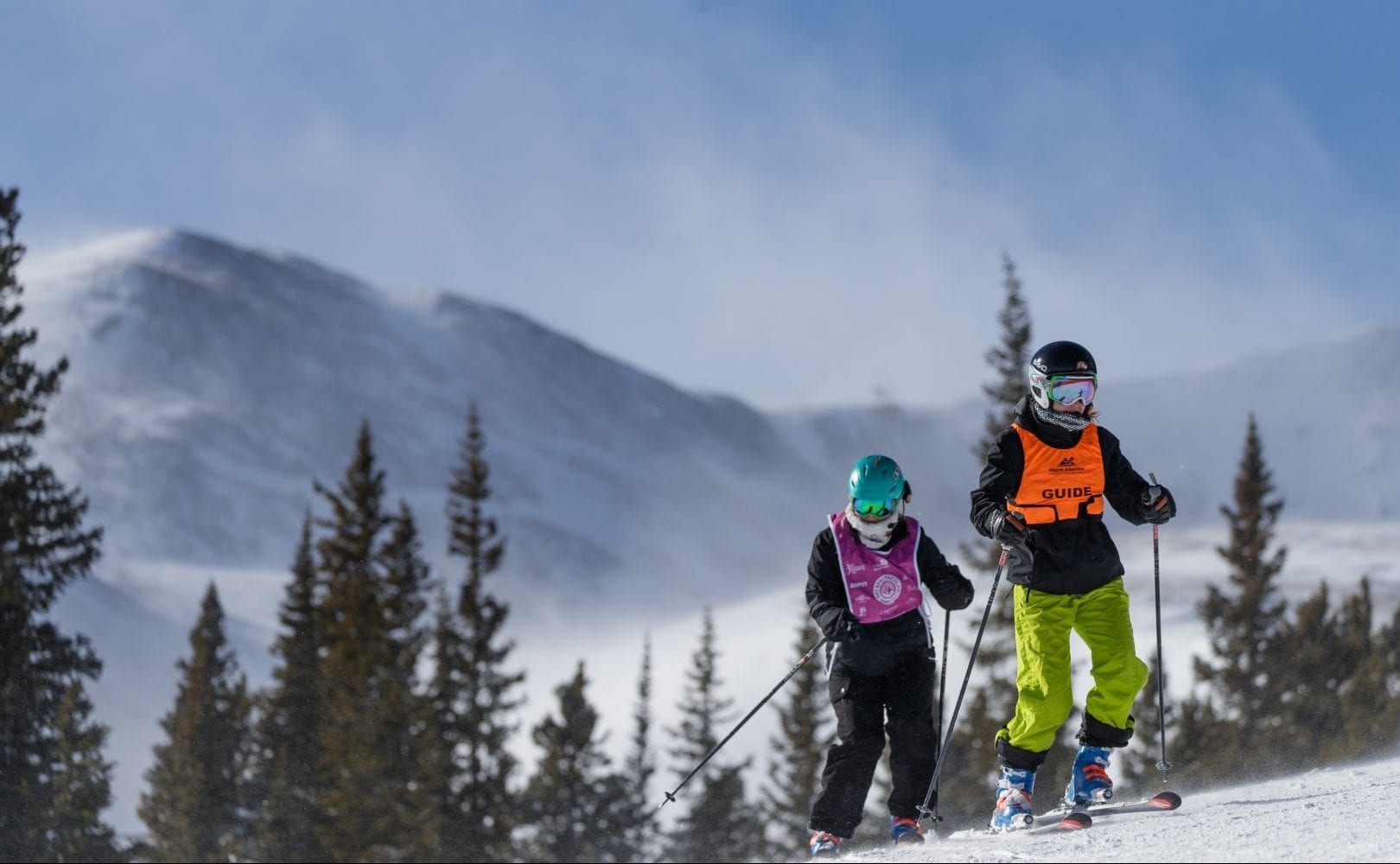 Two skiers on a hill side surrounded by pine trees. In the far distance are snow covered mountains.