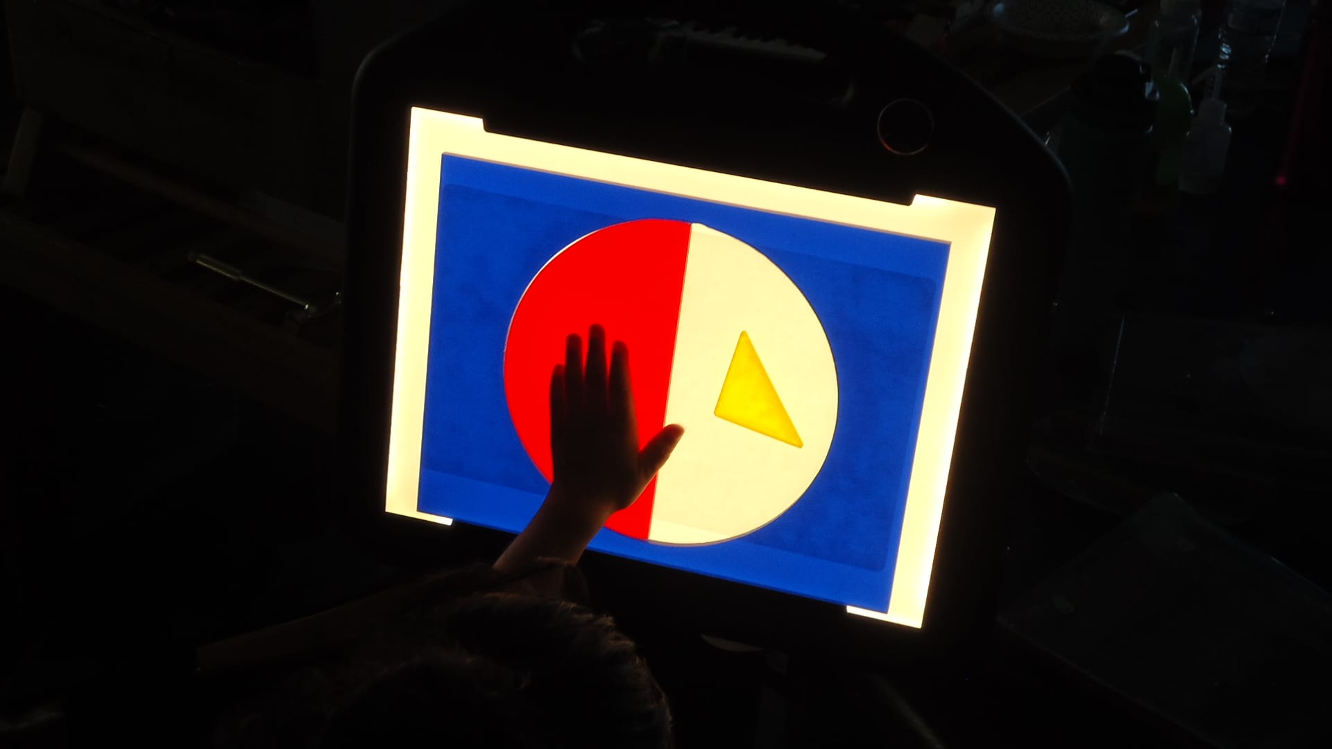 In a darkened room, a child’s hand is touching blue plexiglass frame with a circle cut out. In the circle is a red plexiglass semi-circle and yellow triangle. The black case of the LED Mini-Lite Box disappears into the darkness.