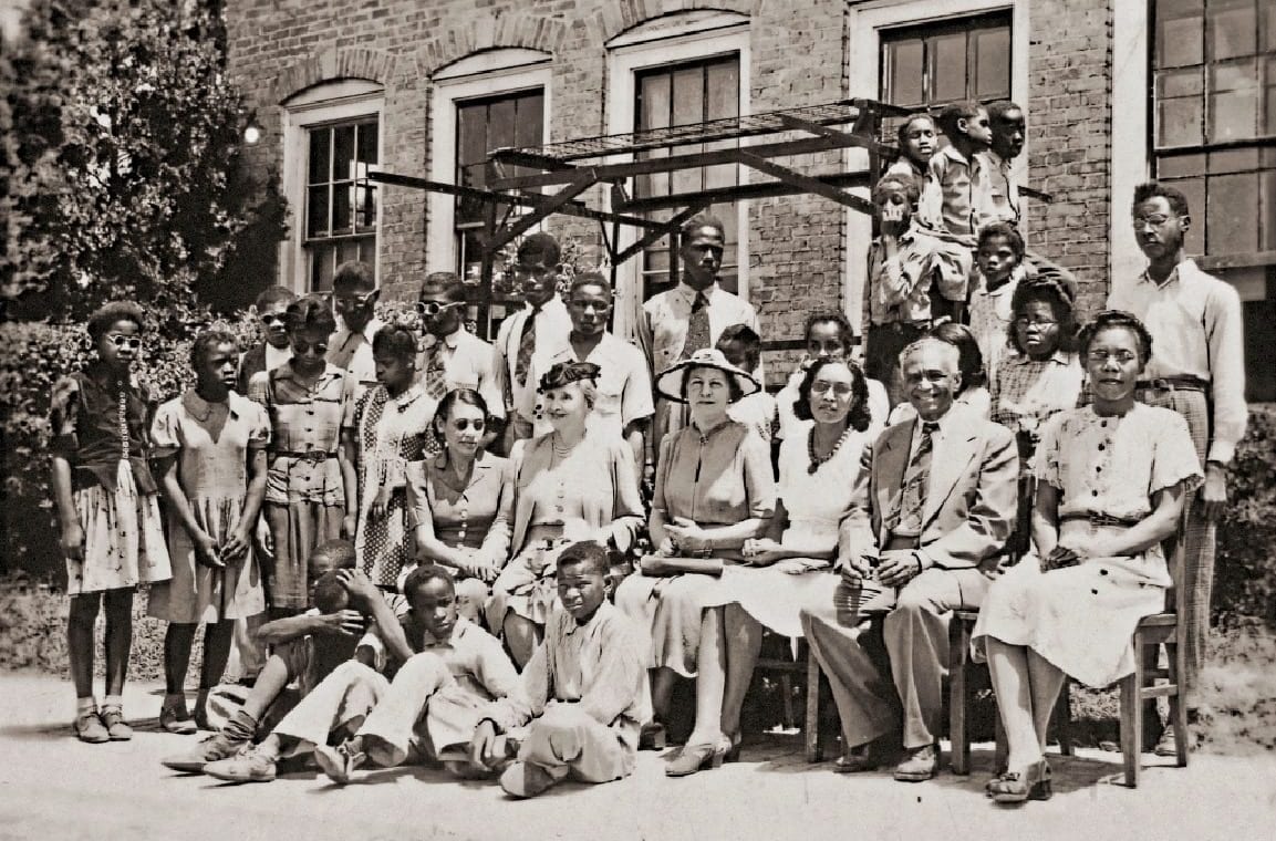 A group of Piney Woods School students and teachers in front of a brick building, with Helen Keller and her assistant Polly Thomson seated in the middle. Martha Foxx sits in the second row on the far left, wearing a light dress with dark buttons, her hands clasped in her lap.