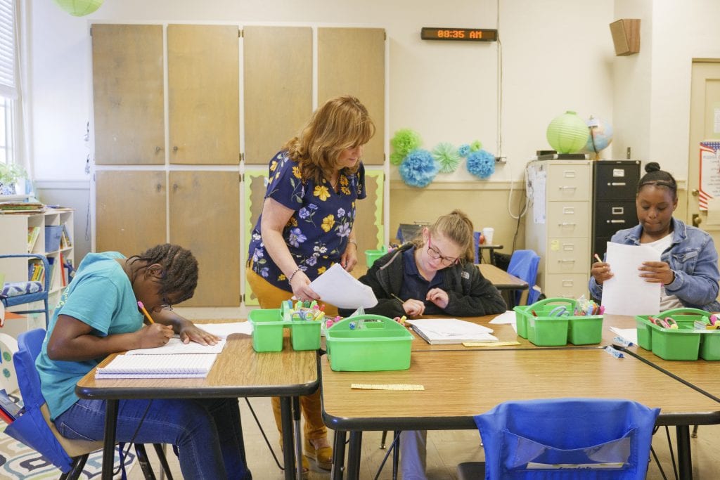 Three students write at a large table in a classroom. A teacher hands a piece of paper to the student nearest to the middle of the table.