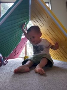 Image: Reach & Match is put up in a configuration like a tent. A little boy who is blind is sitting under and smiling while touching the textures of the Reach & Match mats above him.