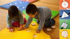 A little girl and little boy are playing on the path side of Reach & Match mats. There are circle, triangle, square, and pentagon tiles that can be put into the corresponding mats.