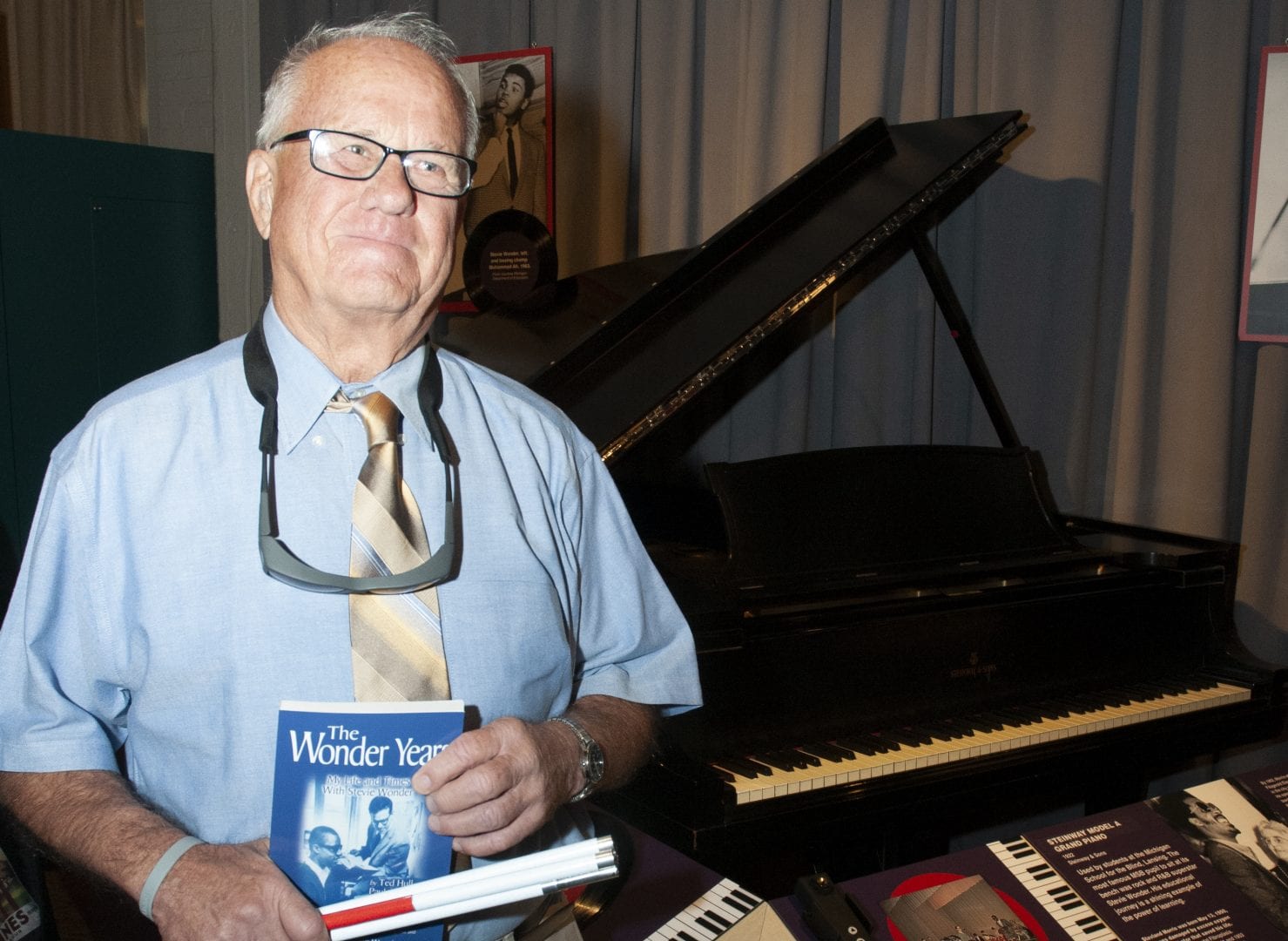 photo of a man standing with a folded white cane in front of a black piano on display in a museum