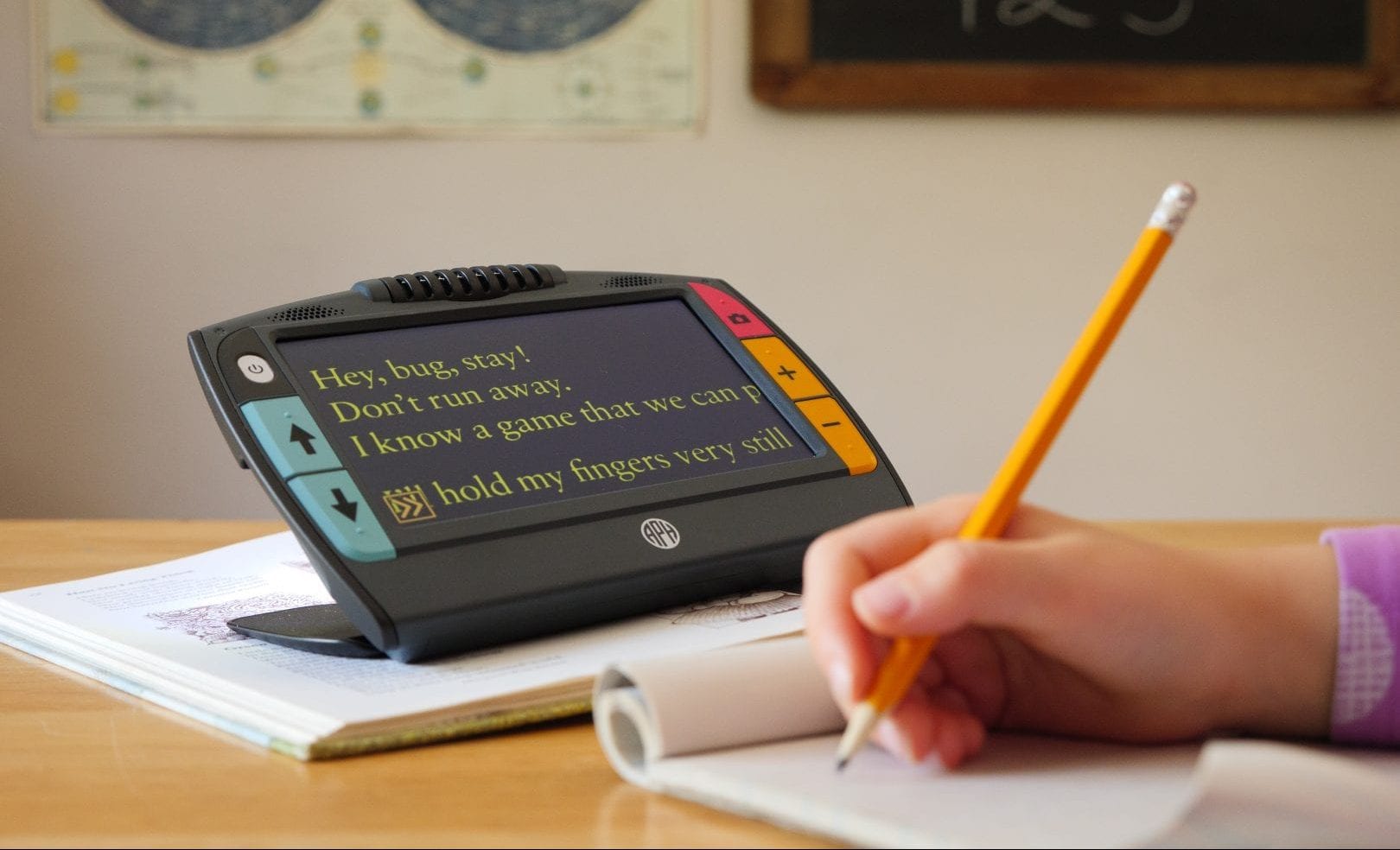 juno magnifier set up on a book magnifying the page at a students desk. student is writing in a notebook with a penil